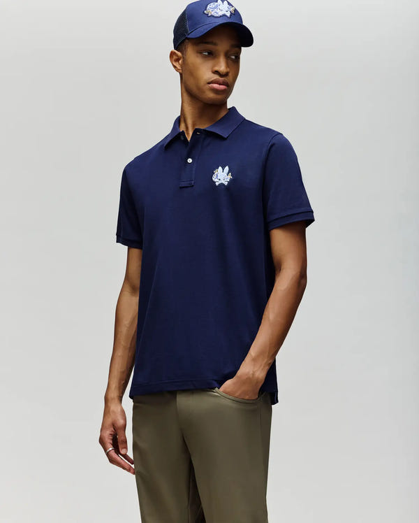 A person wears the Psycho Bunny MENS AUGUSTE PIQUE POLO SHIRT (MT0100051) in navy blue with a white embroidered logo, paired with khaki pants and a matching navy cap featuring the same white logo, against a plain light background.
