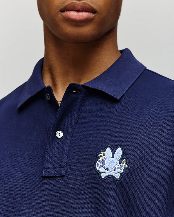 A person wearing the Psycho Bunny MENS AUGUSTE PIQUE POLO SHIRT (MT0100051) in navy blue, featuring a collared neckline, button placket, and a large embroidered rabbit head over crossed bones on the left chest for varsity flair.