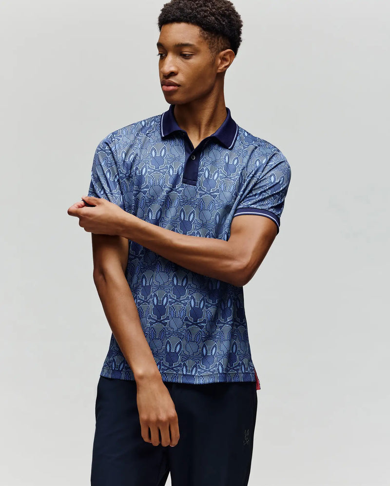 A man in the Psycho Bunny MENS REEVES ALL OVER BUNNY SPORT POLO (MT0100053) and dark trousers stands against a light background, looking left as he adjusts his right sleeve.