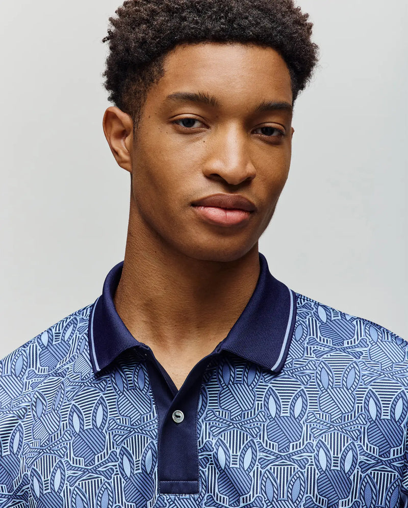 A young man with short curly hair models the Psycho Bunny MENS REEVES ALL OVER BUNNY SPORT POLO (MT0100053), featuring a breathable fabric, subtle blue pattern, and dark blue collar against a plain light background.