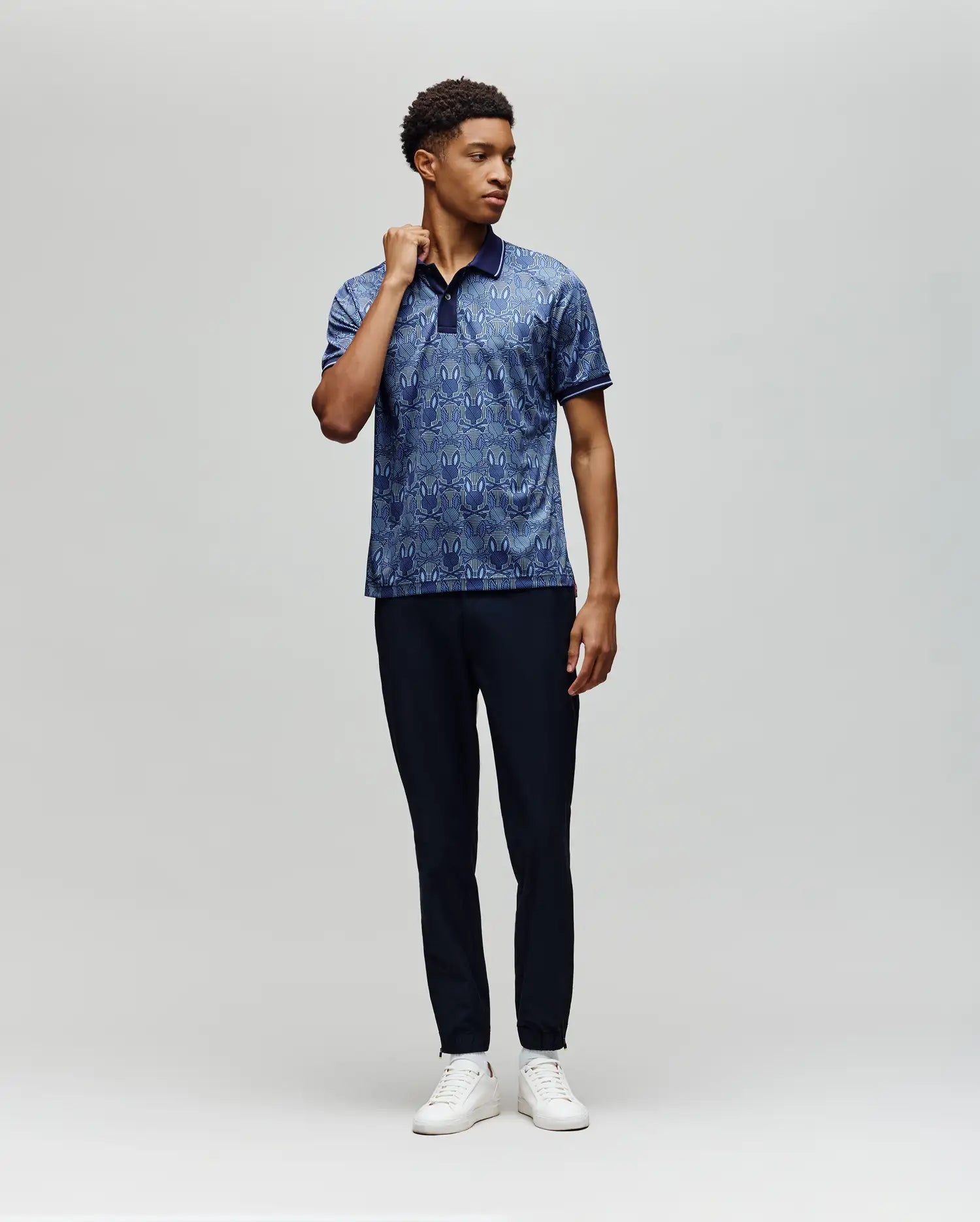 A man stands against a plain light gray background, wearing the Psycho Bunny MENS REEVES ALL OVER BUNNY SPORT POLO (MT0100053), dark navy pants, and white sneakers. He is turned slightly and holds his shirt collar with one hand.