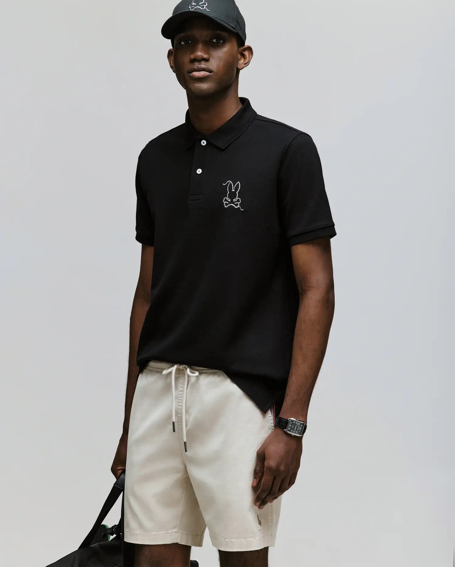 A man in a black Psycho Bunny MENS CYRUS PIQUE POLO (MT0100056), light beige drawstring shorts, and a black baseball cap stands with a black bag and wristwatch against a light gray background.