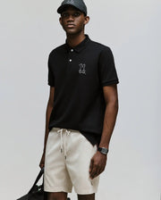 A man in a black Psycho Bunny MENS CYRUS PIQUE POLO (MT0100056), light beige drawstring shorts, and a black baseball cap stands with a black bag and wristwatch against a light gray background.