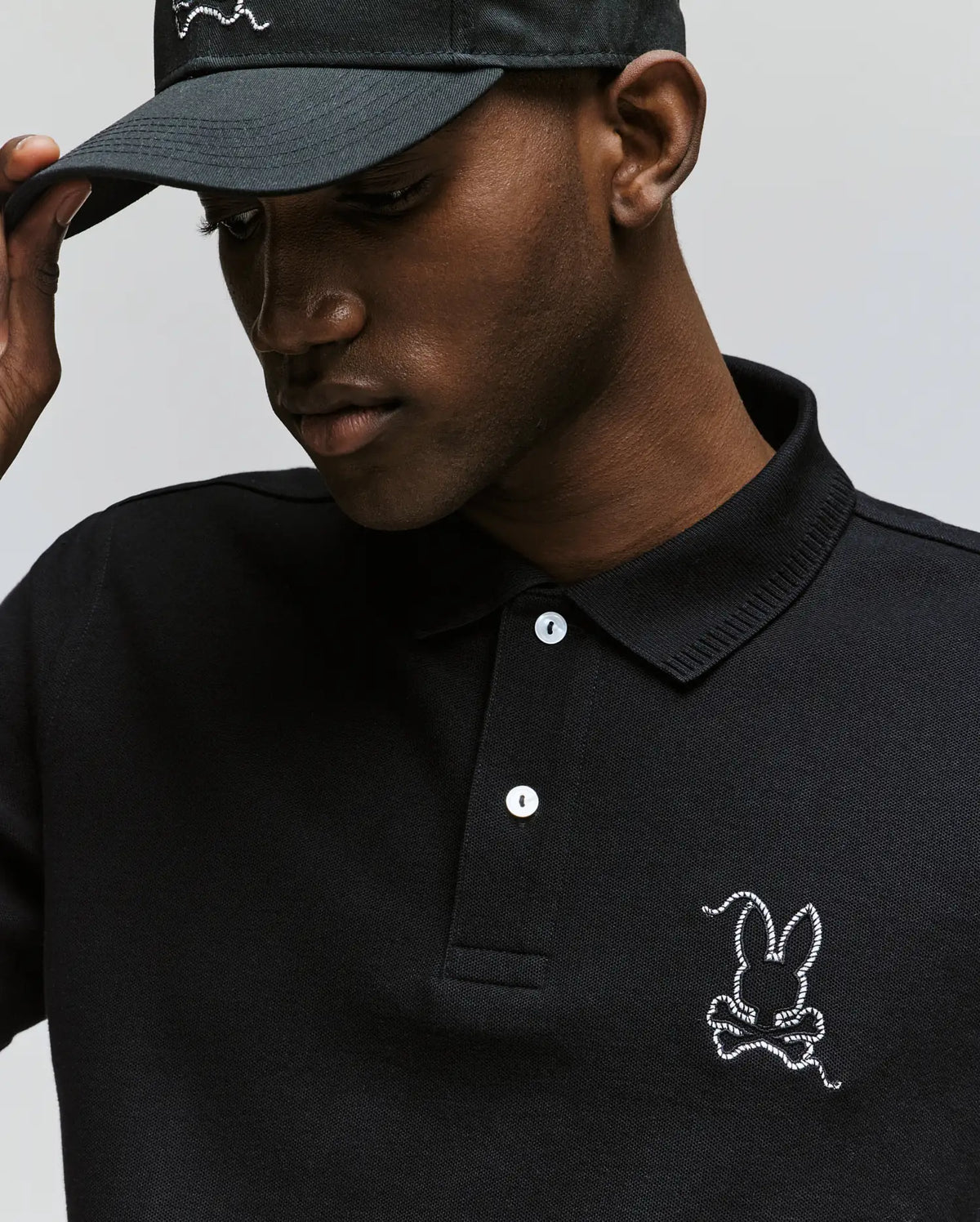 A man in a Psycho Bunny MENS CYRUS PIQUE POLO - MT0100056, featuring soft black Pima cotton and a white bunny logo, adjusts his matching black cap while looking down.