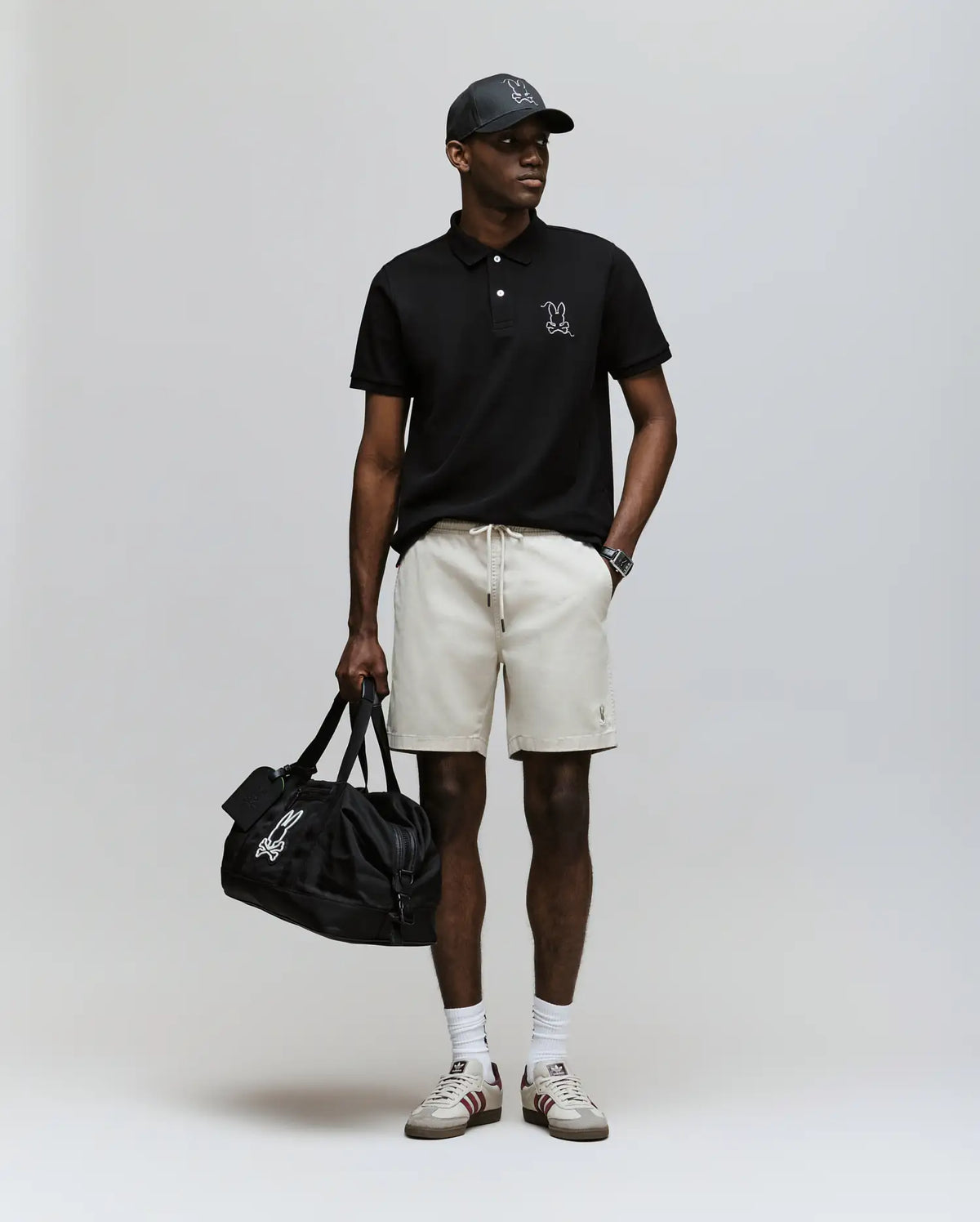 A man wearing a Psycho Bunny MENS CYRUS PIQUE POLO - MT0100056 in black, beige shorts, white socks, sneakers, a black cap, and holding a black duffel bag stands against a plain light background.