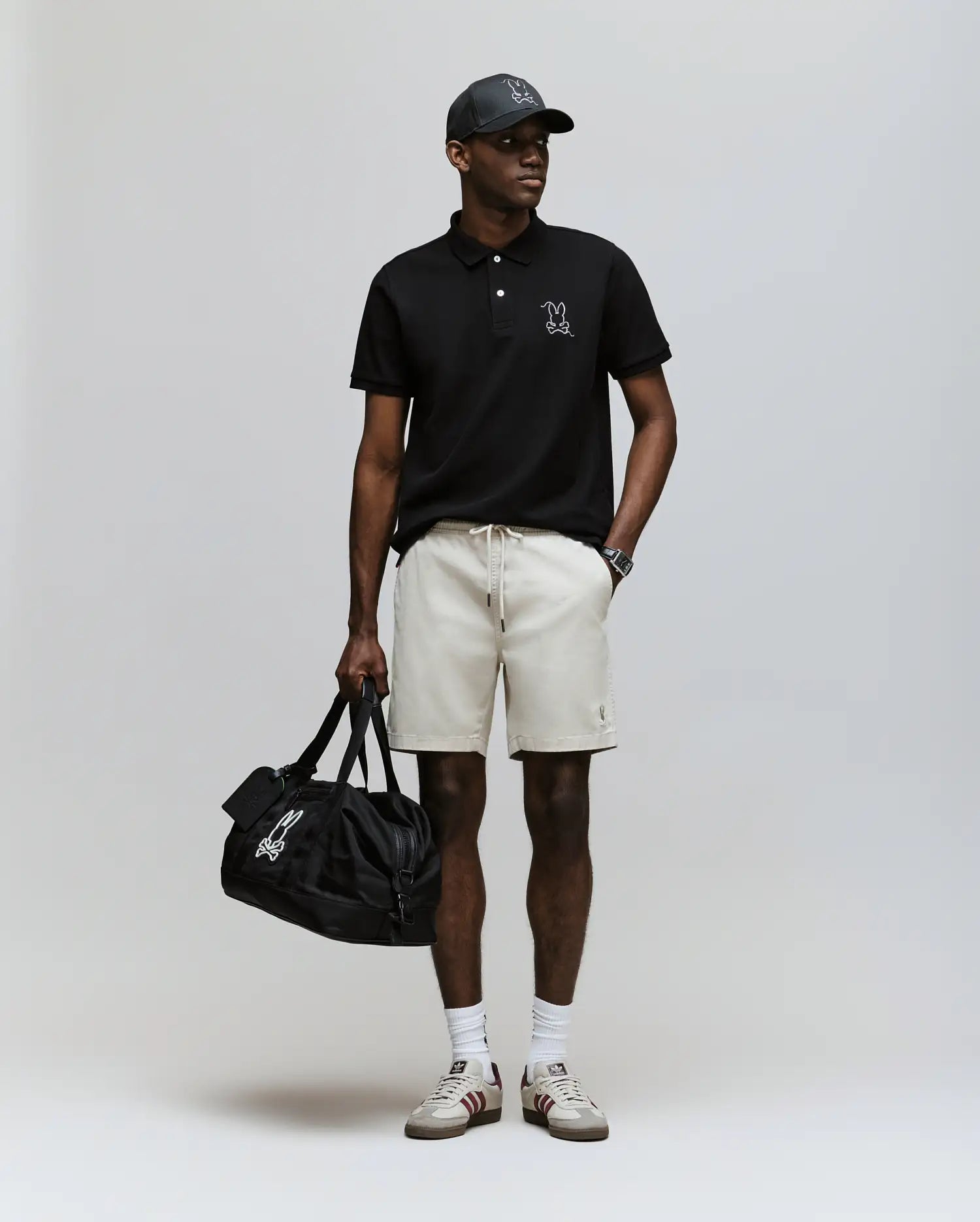 A man wearing a Psycho Bunny MENS CYRUS PIQUE POLO - MT0100056 in black, beige shorts, white socks, sneakers, a black cap, and holding a black duffel bag stands against a plain light background.