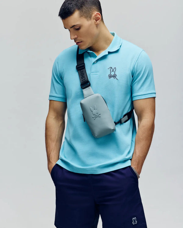 A man in a light blue Psycho Bunny MENS CYRUS PIQUE POLO (MT0100056) with a bunny logo, navy shorts, and a matching light gray crossbody bag stands against a plain background, looking down with one hand in his pocket.