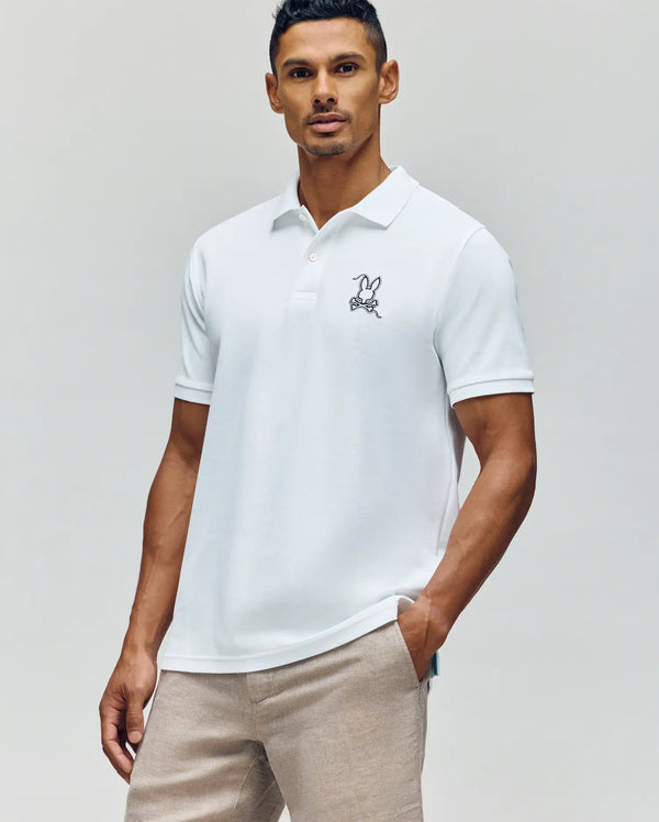 A man wears the Psycho Bunny MENS CYRUS PIQUE POLO - MT0100056 in white, featuring subtle Bunny embroidery on the chest, paired with light beige pants, against a plain light background.
