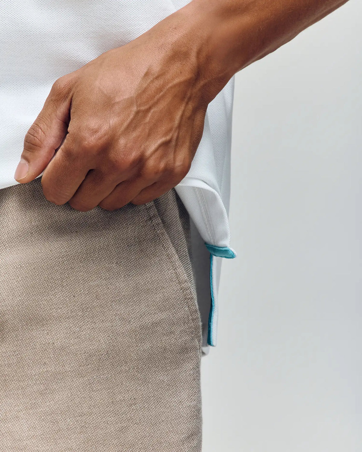 A close-up shows a hand holding the hem of the Psycho Bunny MENS CYRUS PIQUE POLO - MT0100056, white Pima cotton with a small blue tag. The person wears light beige pants against a plain, neutral background.