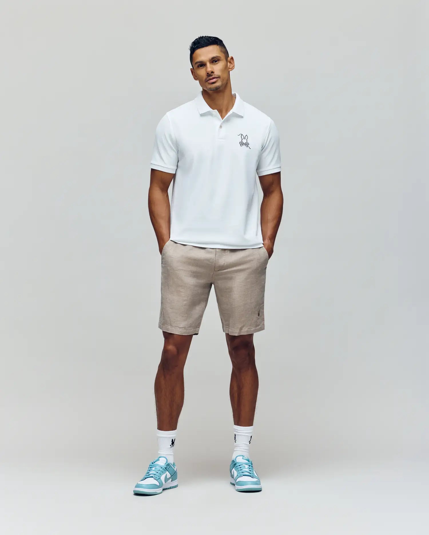 A man stands against a plain background wearing the Psycho Bunny MENS CYRUS PIQUE POLO - MT0100056 in white, beige shorts, white socks, and light blue sneakers. His hands are in his pockets as he looks confidently at the camera.