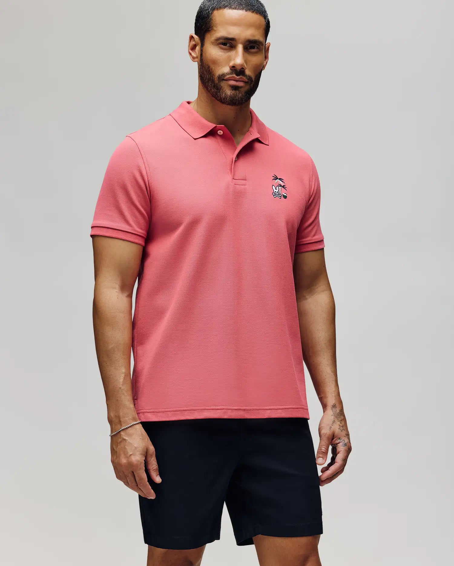 A man with short dark hair and a beard wears the Psycho Bunny MENS SONNY PIQUE POLO - MT0100094 in pink, featuring chest embroidery, paired with black shorts, standing against a plain light gray background.