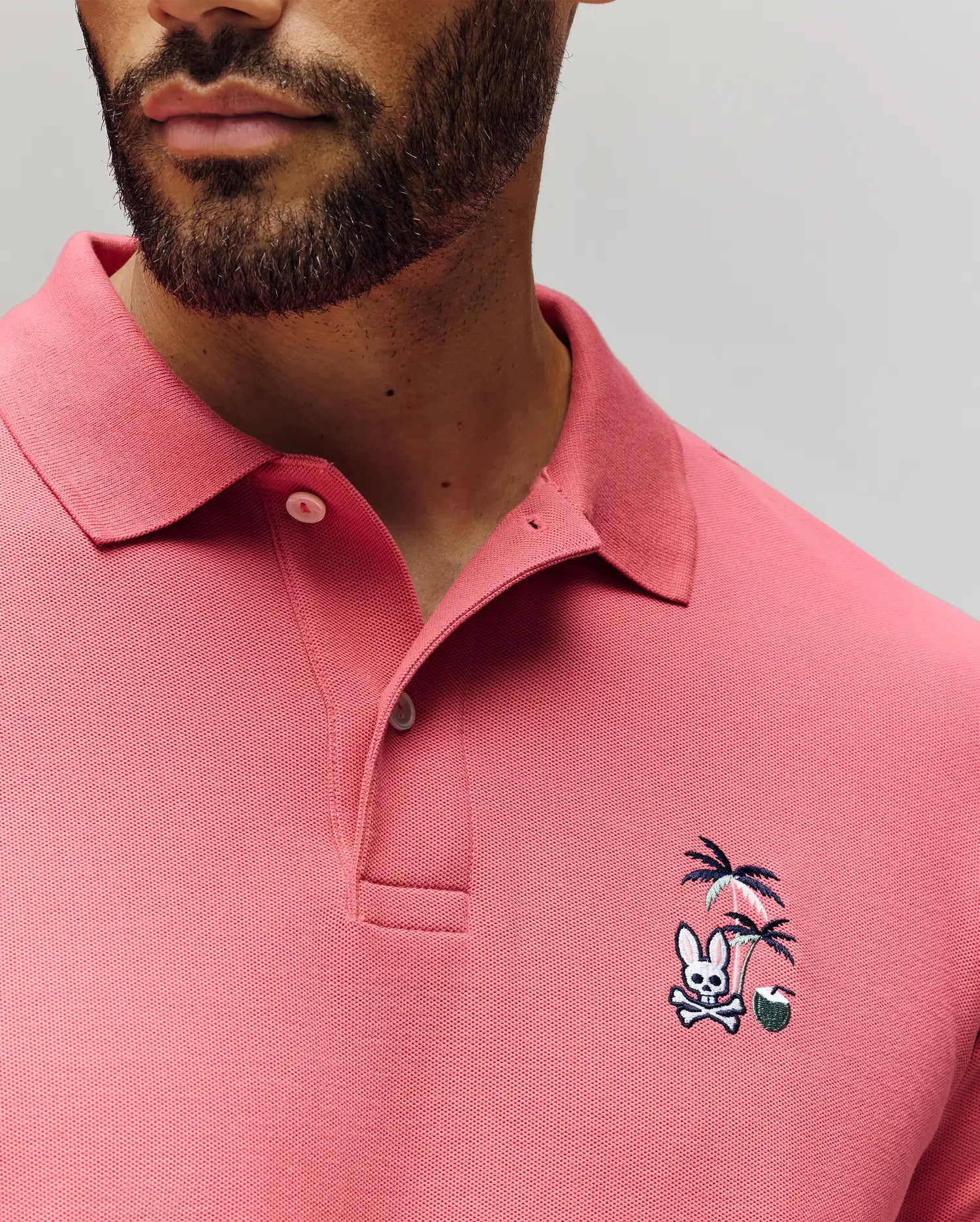 A man wears the Psycho Bunny MENS SONNY PIQUE POLO—crafted from soft Pima cotton—featuring unique chest embroidery: a skull and crossbones under palm trees with a small green ball.