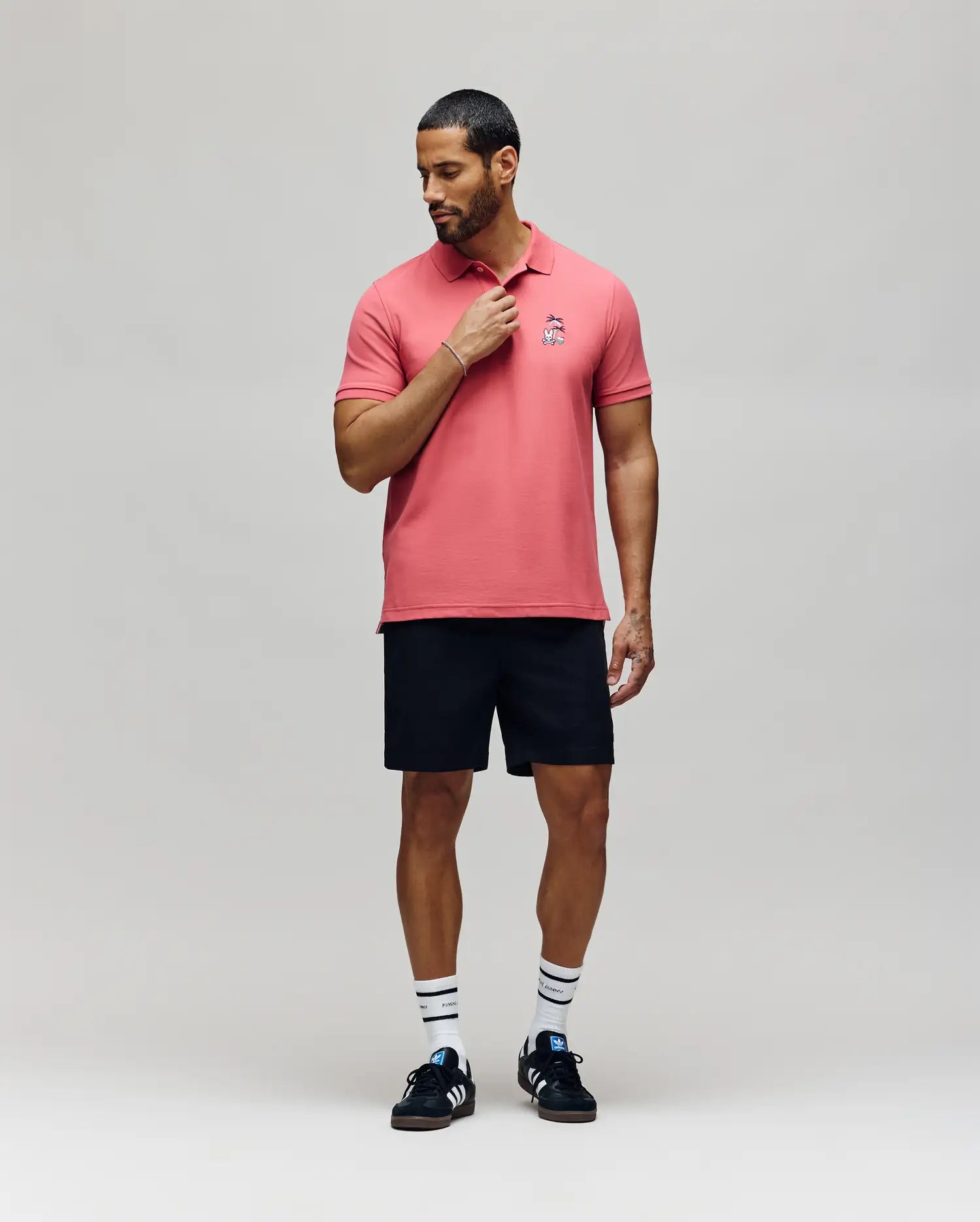 A man with short hair and a beard stands against a plain background, touching his collar as he looks down. He wears Psycho Bunny's MENS SONNY PIQUE POLO - MT0100094 in pink, black shorts, striped white socks, and black sneakers.