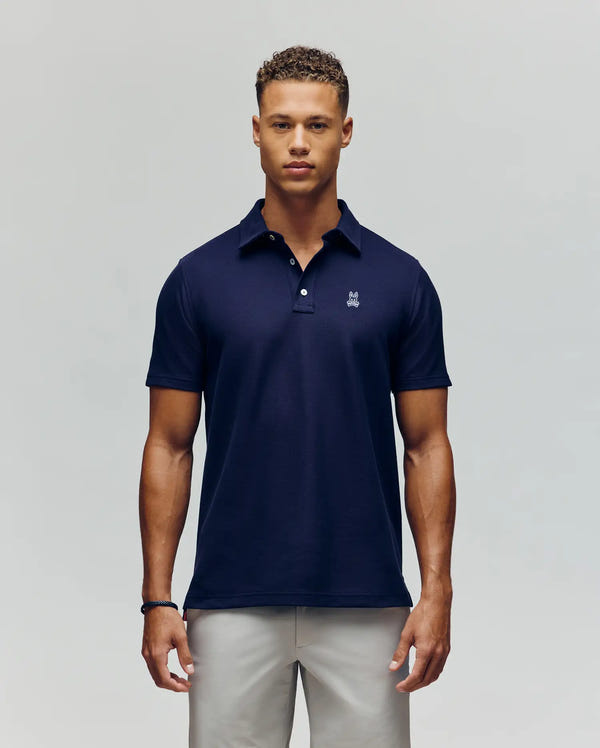 A man faces forward against a plain background, wearing the Psycho Bunny MENS PHOENIX FASHION POLO - MT0100096 in navy blue with a small white logo on the chest and light-colored pants.