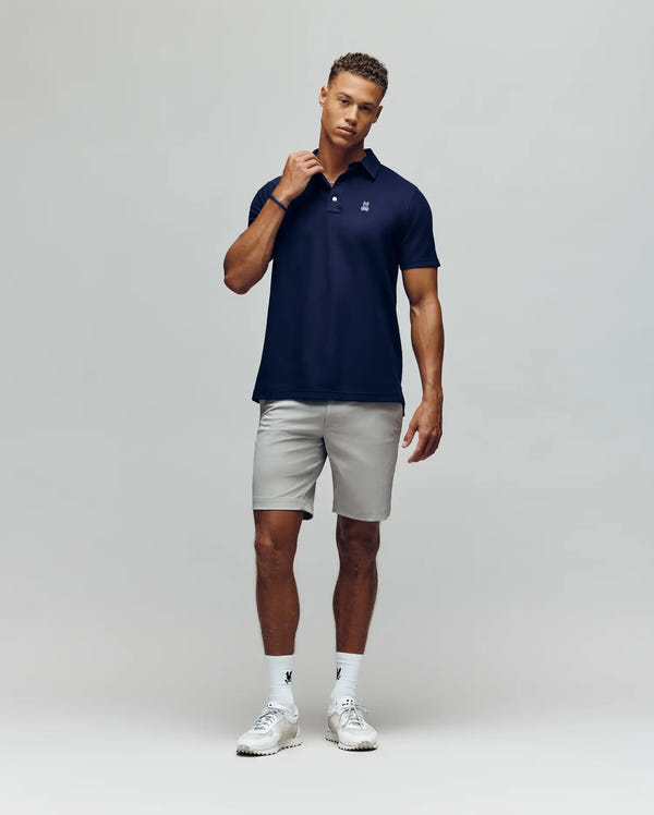 A man stands against a plain background wearing the Psycho Bunny MENS PHOENIX FASHION POLO - MT0100096 in navy, paired with light gray shorts, white athletic shoes, and white socks. He holds the shirt collar and looks calmly at the camera.