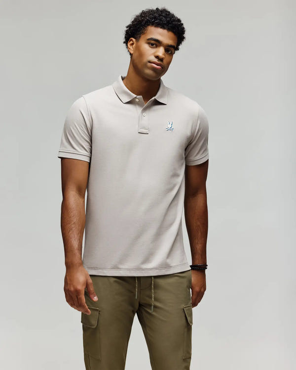 A man stands against a plain background wearing the Psycho Bunny MENS CASSIAN PIQUE POLO - MT0100098, a light beige short-sleeve Pima cotton shirt with a small chest logo, and olive green cargo pants, arms relaxed by his sides.