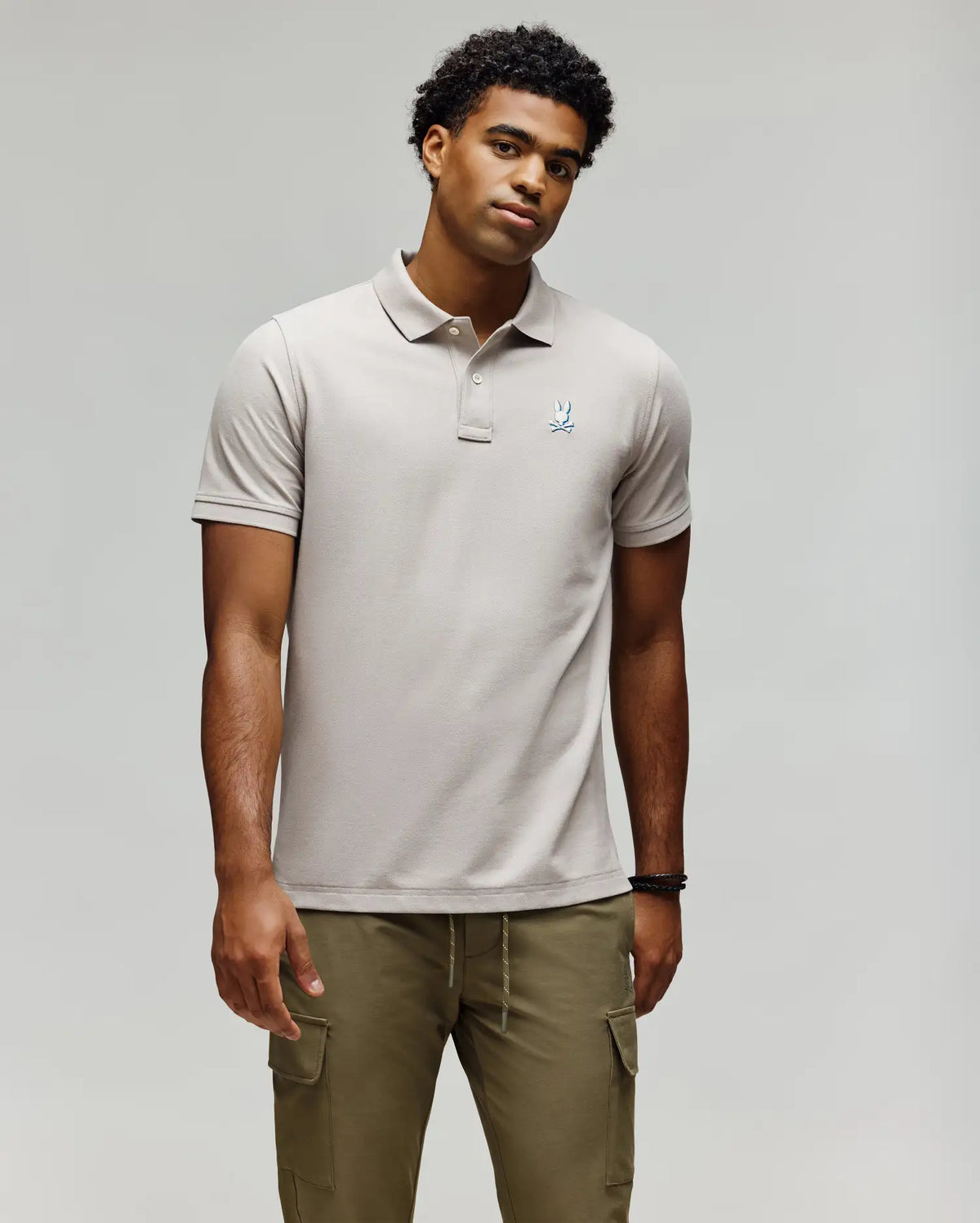 A man stands against a plain background wearing the Psycho Bunny MENS CASSIAN PIQUE POLO - MT0100098, a light beige short-sleeve Pima cotton shirt with a small chest logo, and olive green cargo pants, arms relaxed by his sides.