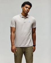 A man stands against a plain background wearing the Psycho Bunny MENS CASSIAN PIQUE POLO - MT0100098, a light beige short-sleeve Pima cotton shirt with a small chest logo, and olive green cargo pants, arms relaxed by his sides.