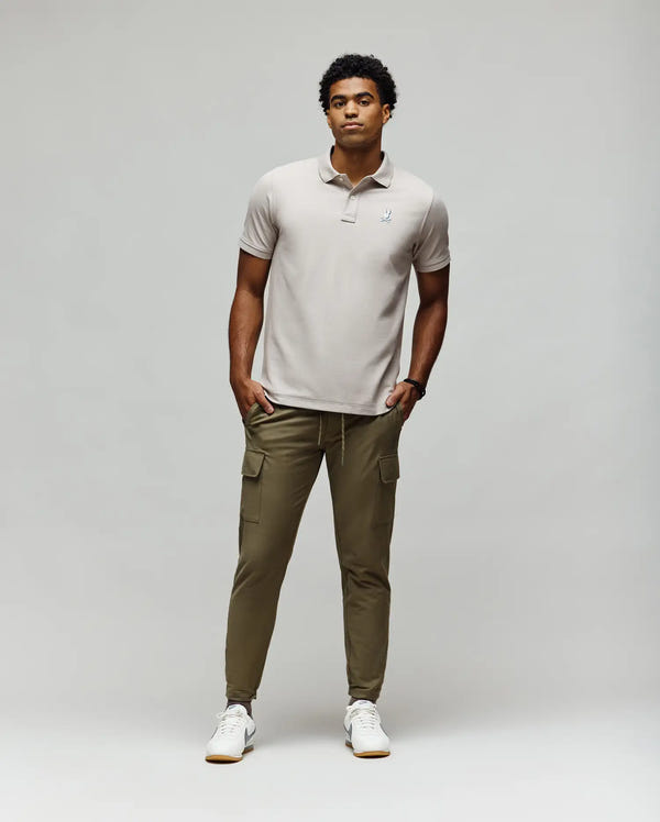 A man stands against a plain light background wearing the Psycho Bunny MENS CASSIAN PIQUE POLO - MT0100098 in light gray, olive green cargo joggers, and white sneakers. His hands are in his pockets as he looks slightly to the side.