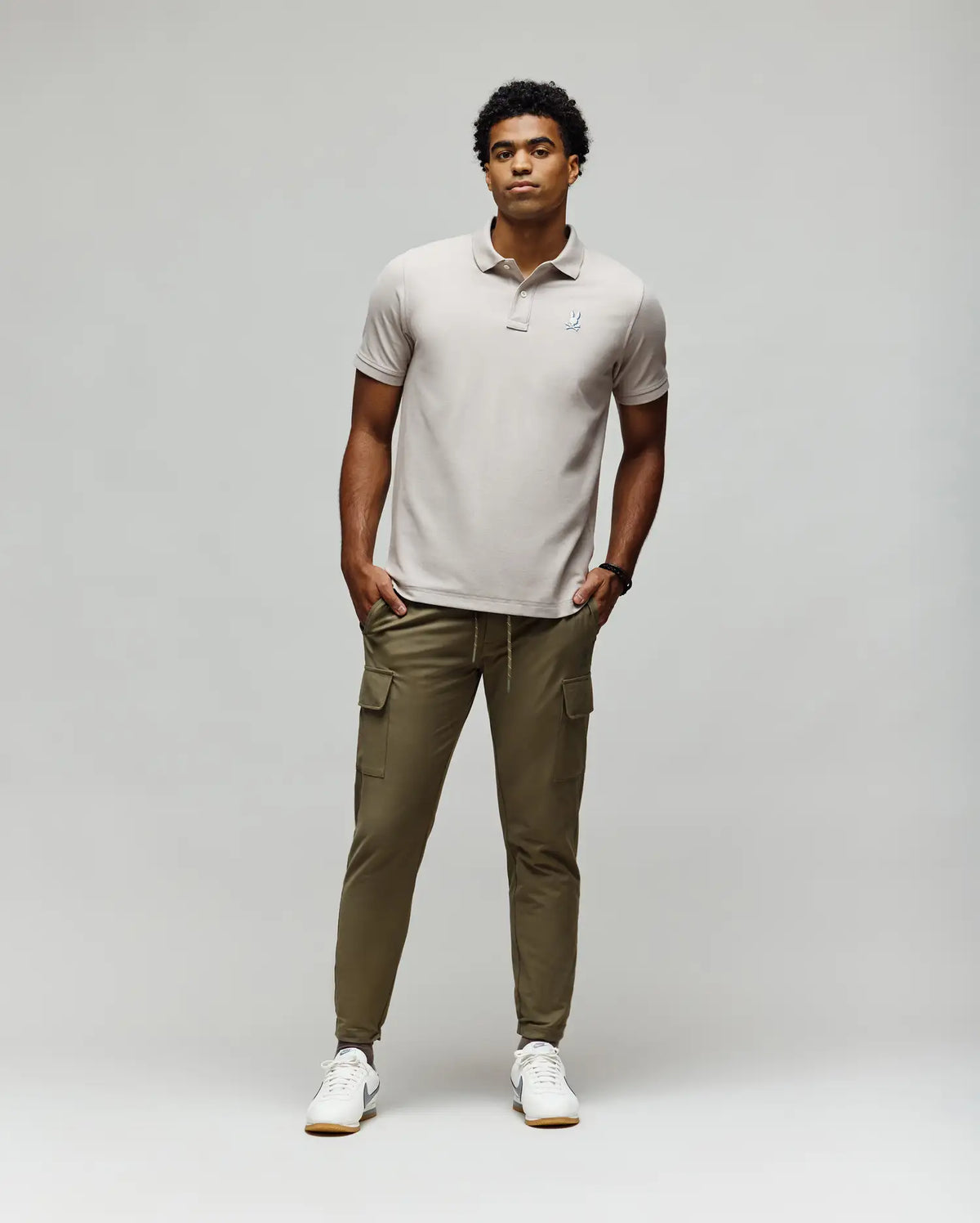 A man stands against a plain light background wearing the Psycho Bunny MENS CASSIAN PIQUE POLO - MT0100098 in light gray, olive green cargo joggers, and white sneakers. His hands are in his pockets as he looks slightly to the side.