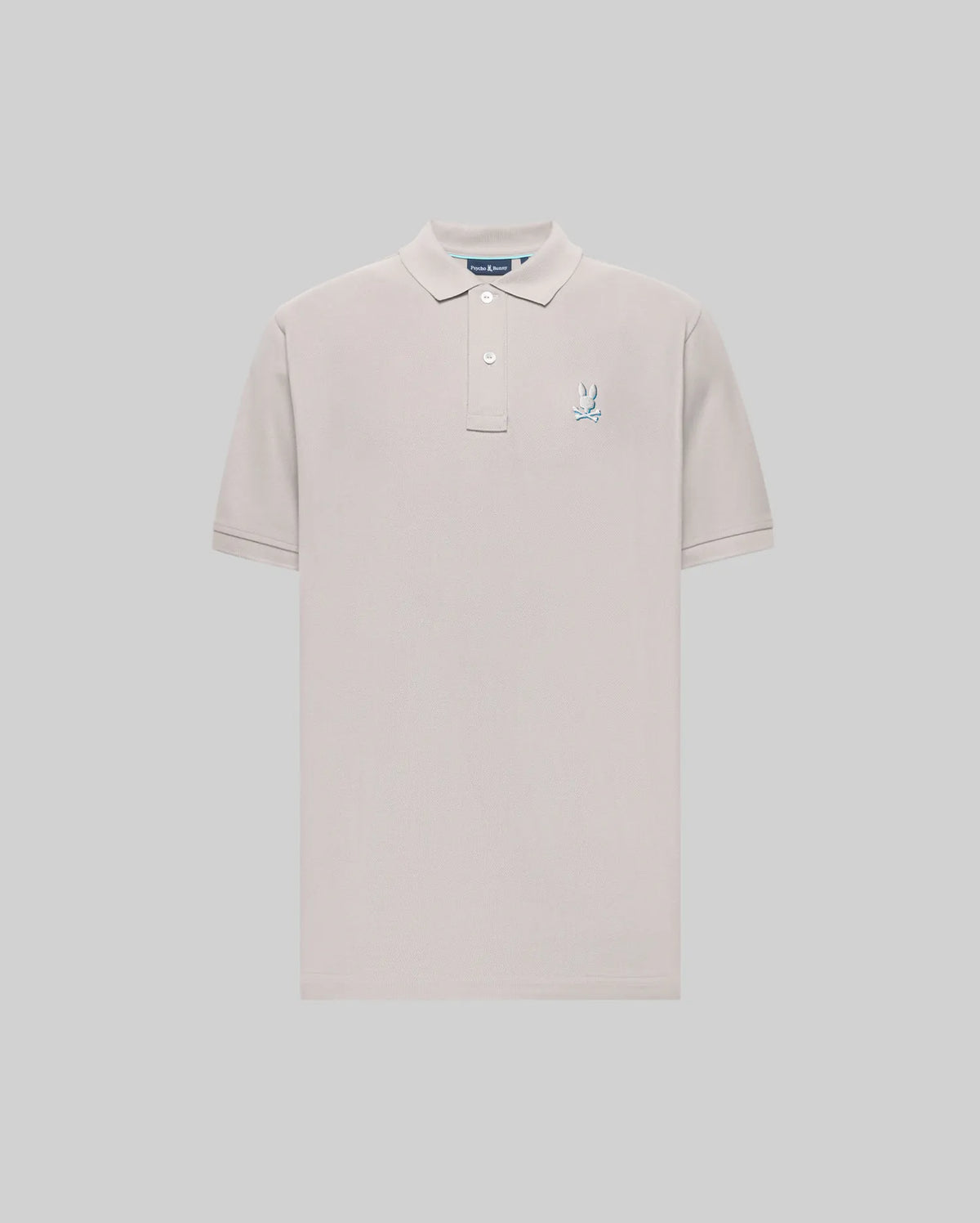 The Psycho Bunny MENS CASSIAN PIQUE POLO (MT0100098) is a light beige, short-sleeve Pima cotton shirt with a collar, two buttons, and a blue and white embroidered bunny logo on the left chest, shown on a plain gray background.