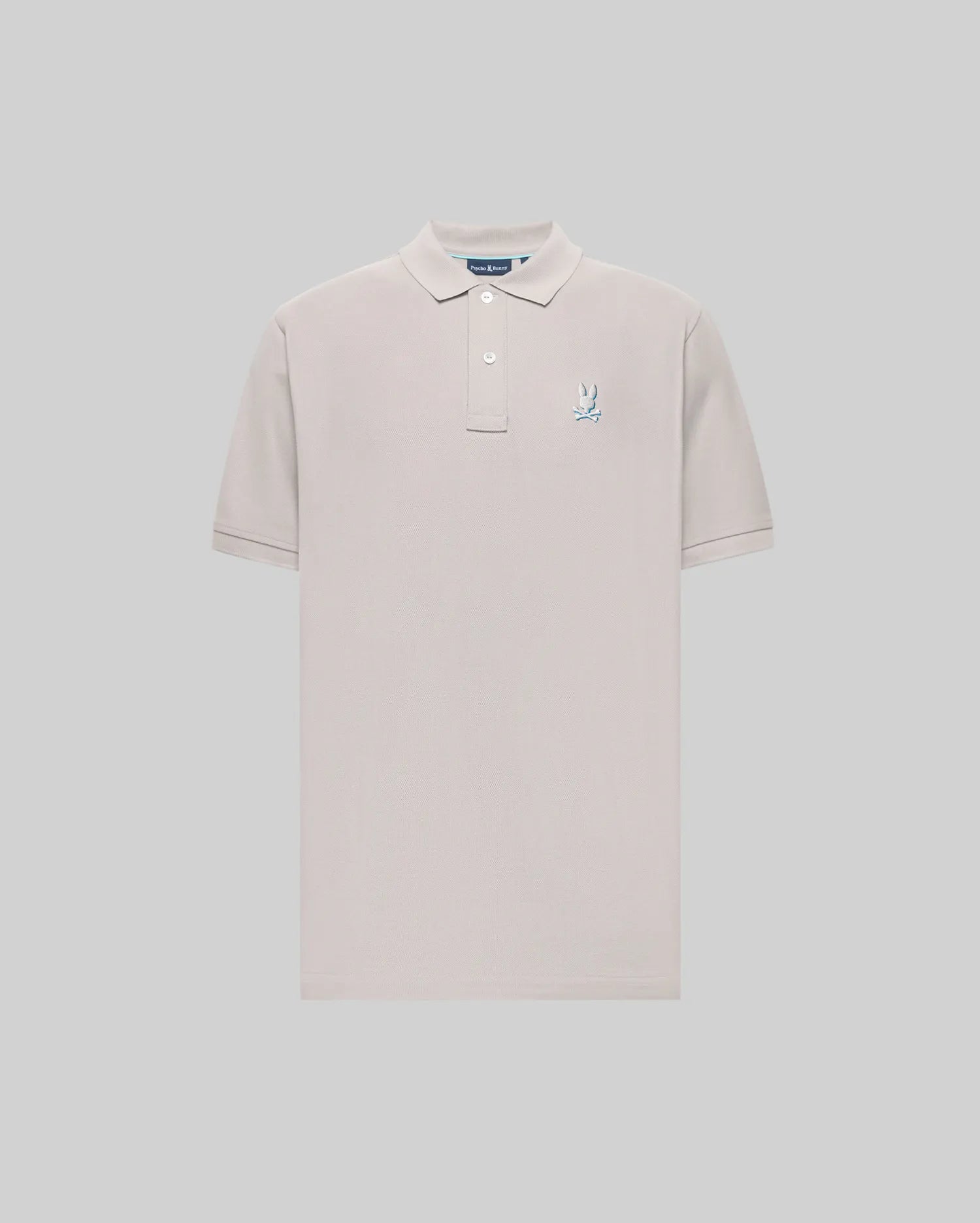 The Psycho Bunny MENS CASSIAN PIQUE POLO (MT0100098) is a light beige, short-sleeve Pima cotton shirt with a collar, two buttons, and a blue and white embroidered bunny logo on the left chest, shown on a plain gray background.