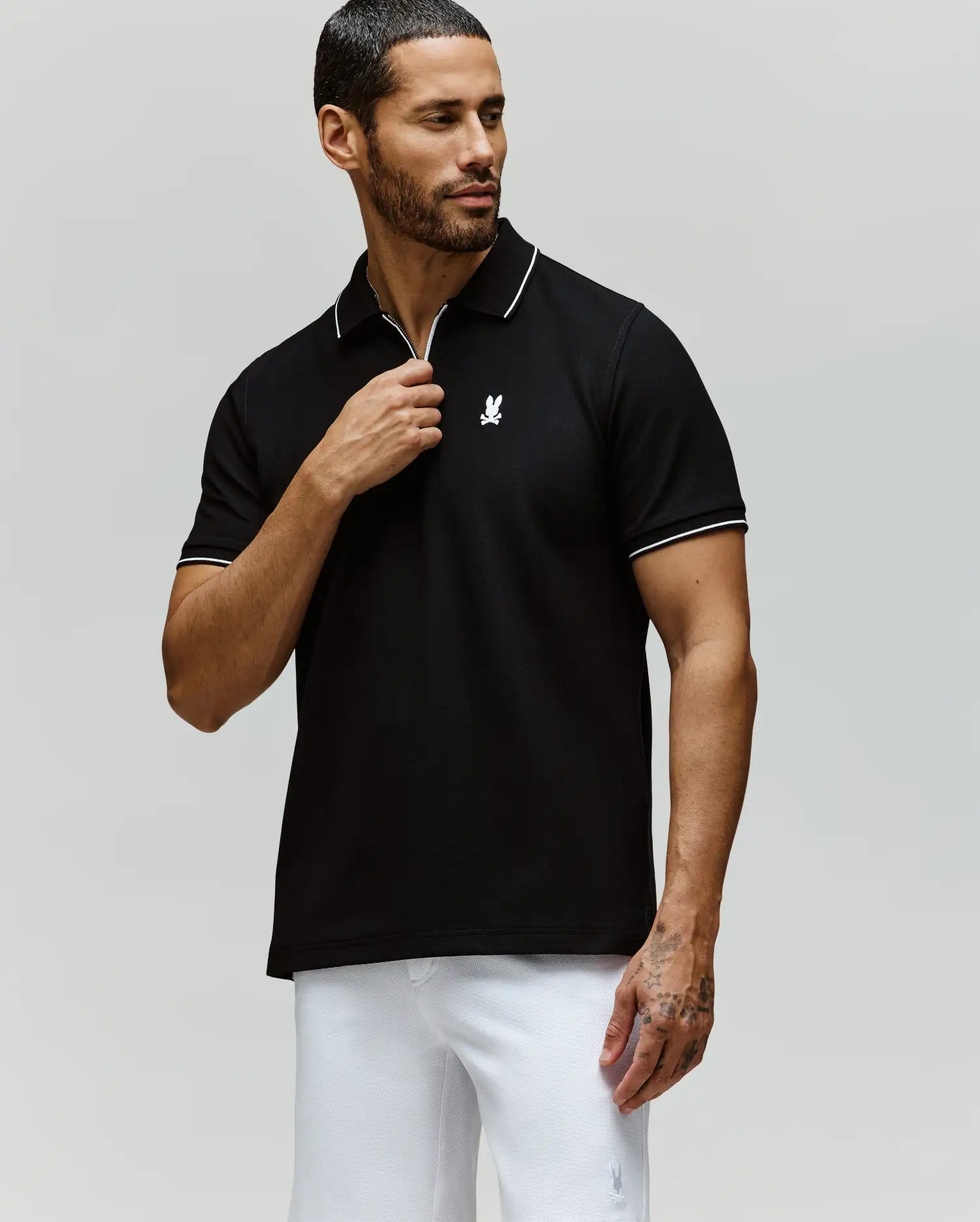 A man with short dark hair and a beard wears the Psycho Bunny MENS DANE ZIP PIQUE POLO - MT0100103 in black with white trim and an embroidered Bunny logo, paired with white pants, standing against a plain light grey background.