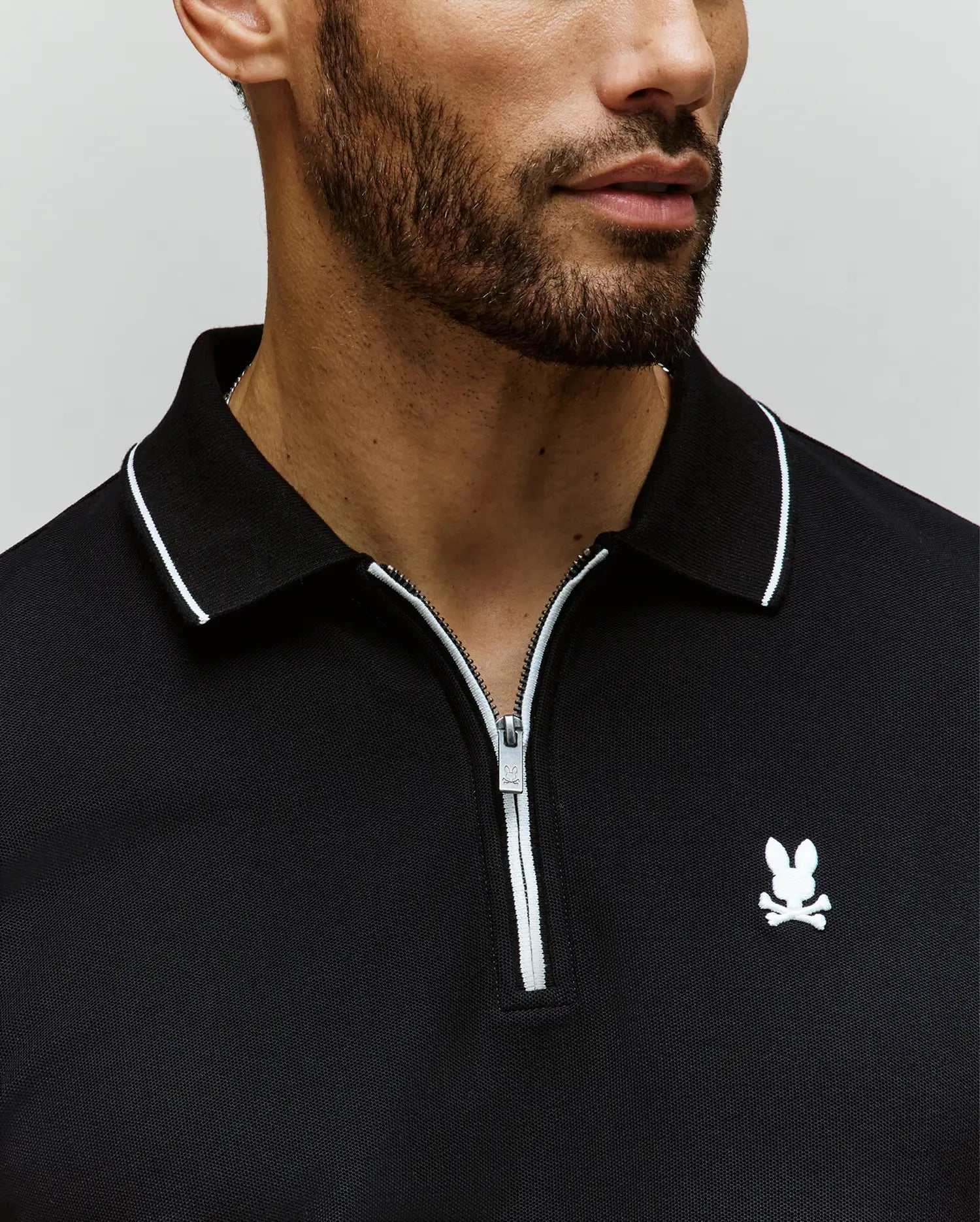 A man with a short beard wears the Psycho Bunny MENS DANE ZIP PIQUE POLO (MT0100103), a black collared zip-up shirt with white trim, sleek zip placket, and an embroidered Bunny logo on the chest. Image is cropped at nose and shoulders.