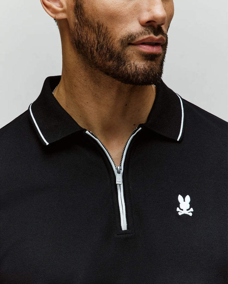 A man with a short beard wears the Psycho Bunny MENS DANE ZIP PIQUE POLO (MT0100103), a black collared zip-up shirt with white trim, sleek zip placket, and an embroidered Bunny logo on the chest. Image is cropped at nose and shoulders.