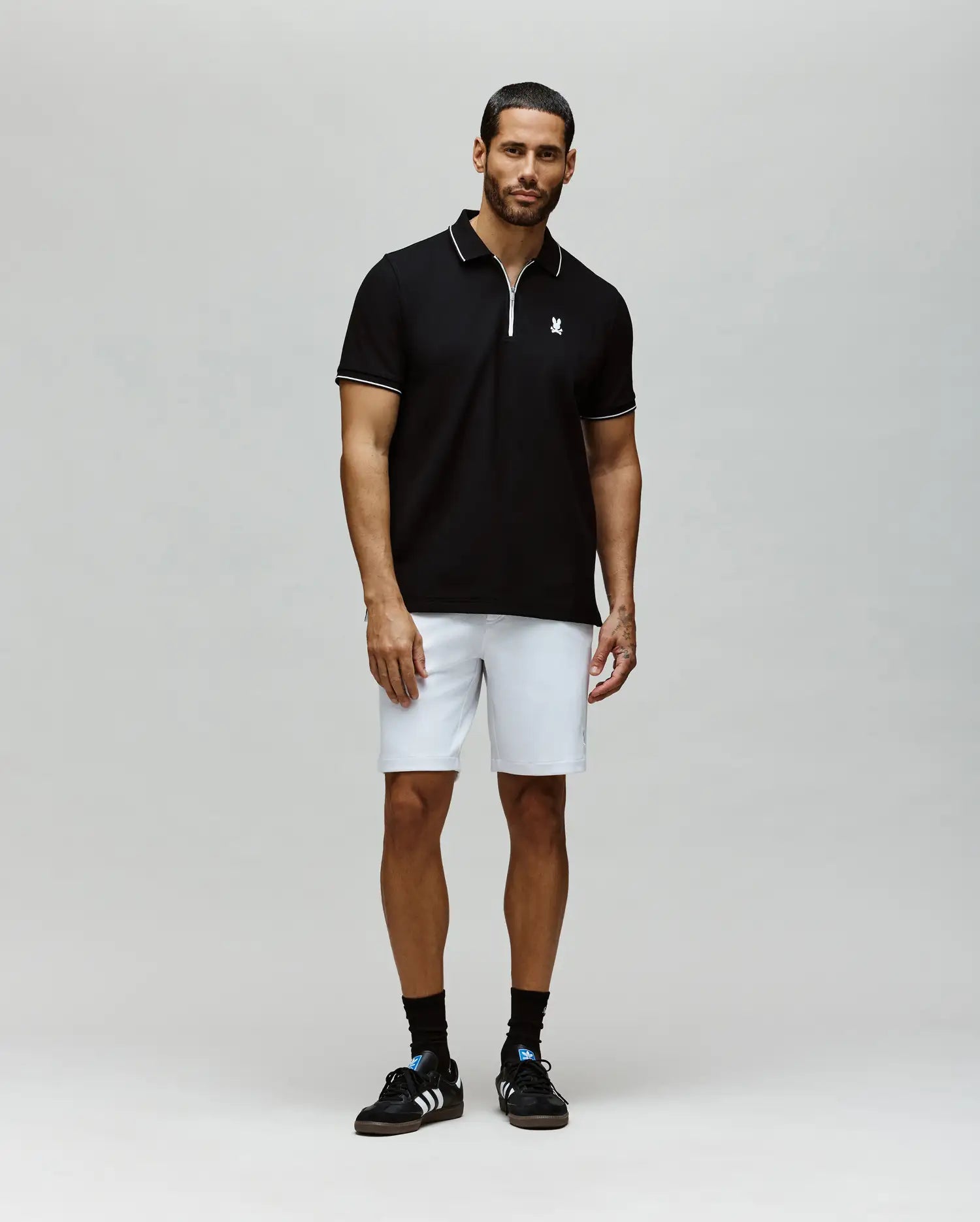 A man stands against a plain background wearing the Psycho Bunny MENS DANE ZIP PIQUE POLO - MT0100103 in black with white trim, paired with white knee-length shorts, black crew socks, and black sneakers.
