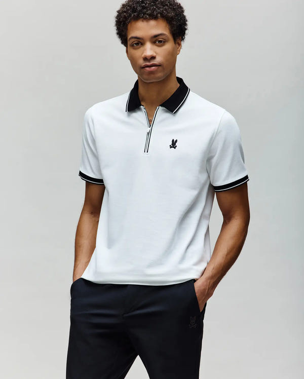 A man wears the Psycho Bunny MENS DANE ZIP PIQUE POLO (MT0100103), a white Pima cotton short-sleeve shirt with black collar and sleeve trim and an embroidered Bunny, standing with hands in pockets against a plain light background.