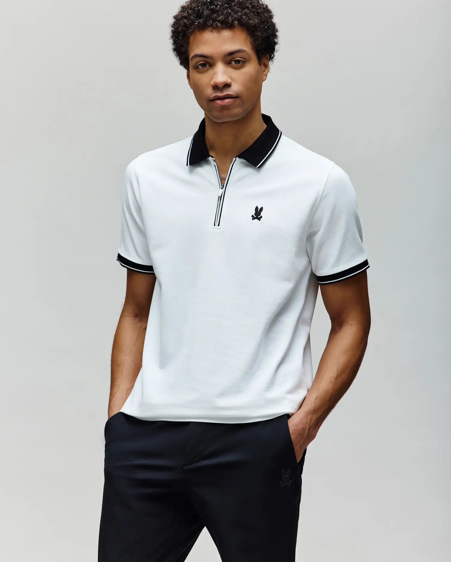 A man wears the Psycho Bunny MENS DANE ZIP PIQUE POLO (MT0100103), a white Pima cotton short-sleeve shirt with black collar and sleeve trim and an embroidered Bunny, standing with hands in pockets against a plain light background.