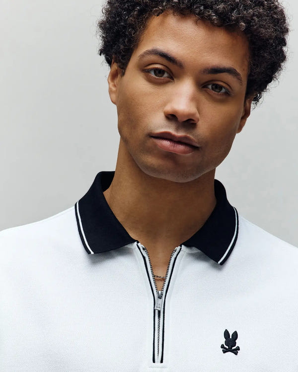 A young man with short curly hair wears the Psycho Bunny MENS DANE ZIP PIQUE POLO (MT0100103), a regular fit white collared shirt with black trim and zipper, facing the camera against a plain, light background.