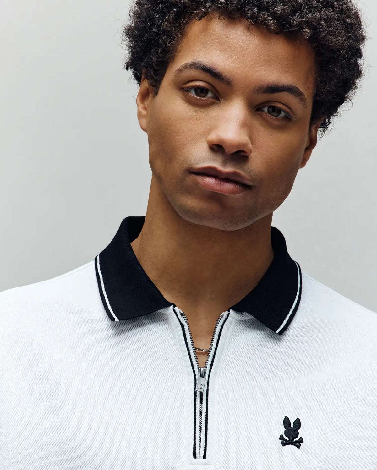 A young man with short curly hair wears the Psycho Bunny MENS DANE ZIP PIQUE POLO (MT0100103), a regular fit white collared shirt with black trim and zipper, facing the camera against a plain, light background.