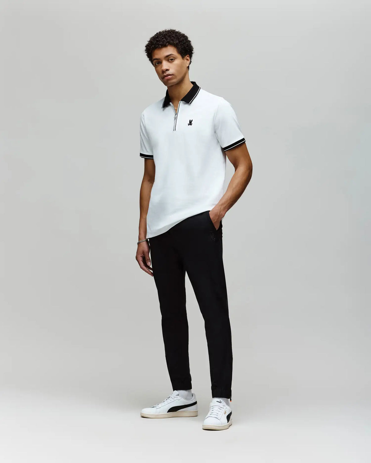 A young man with short curly hair stands against a plain background, wearing Psycho Bunny’s MENS DANE ZIP PIQUE POLO - MT0100103 in white with black trim and an embroidered bunny, paired with black pants and white sneakers, his posture relaxed.