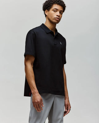 A young man with short curly hair wears the Psycho Bunny MENS HENRY OVERSIZED FIT POLO (MT0100104) in black and light gray pants, standing against a plain light gray background, looking slightly to the side with a neutral expression.