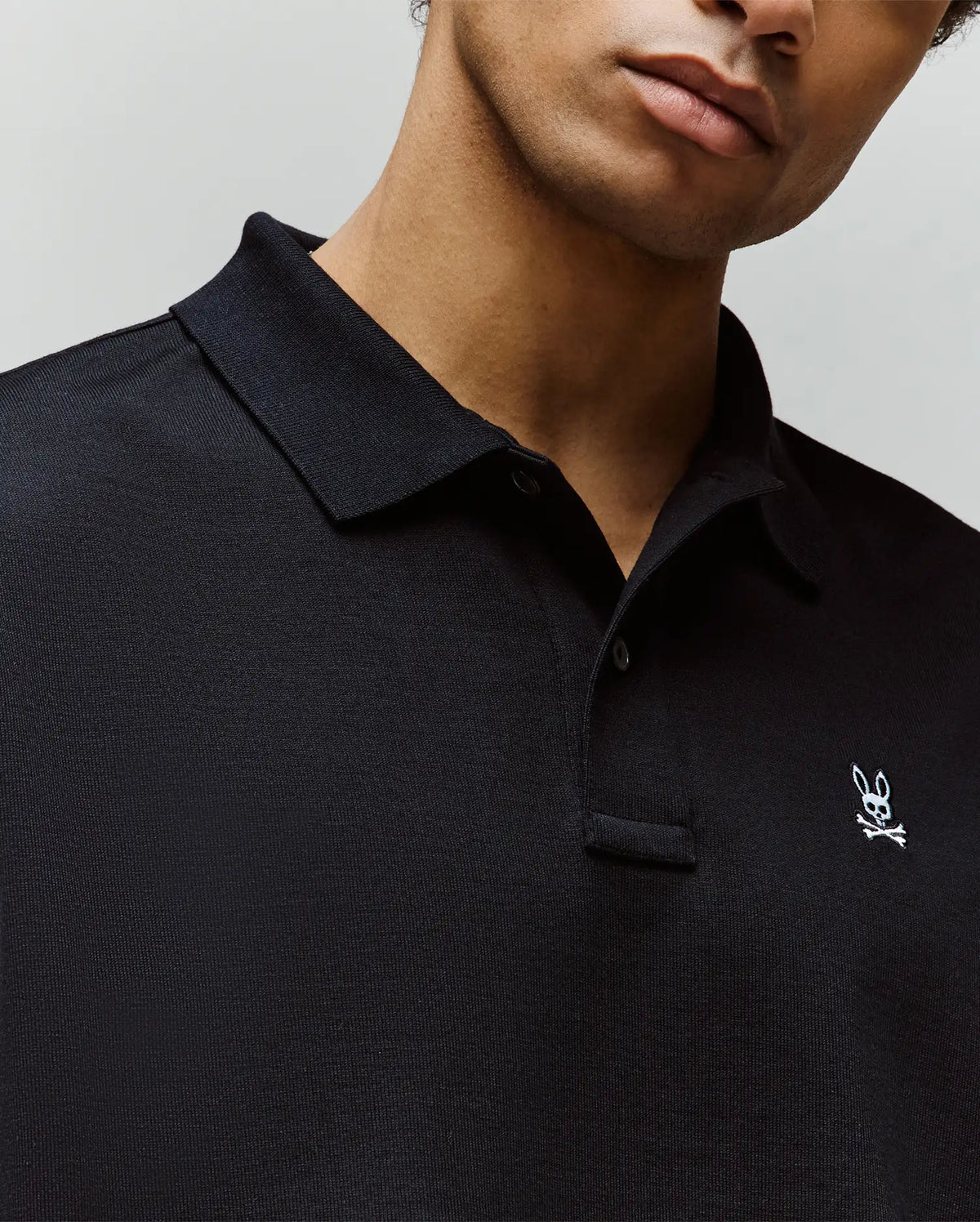 A man in a Psycho Bunny MENS HENRY OVERSIZED FIT POLO (MT0100104) made of heavyweight jersey, featuring a small white skull and bunny ears logo on the chest; only his lower face and upper torso are visible.