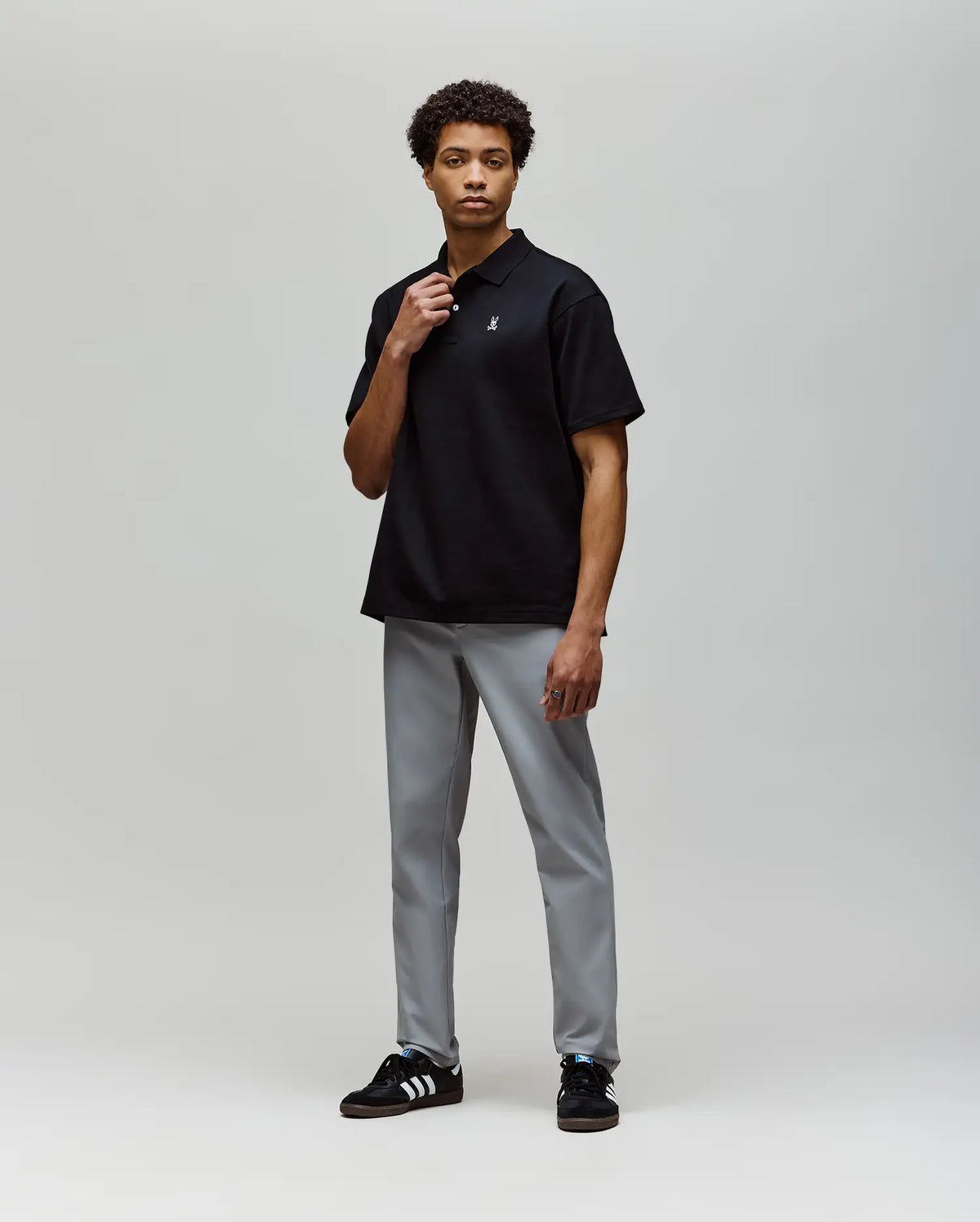 A man stands in a studio against a light gray background, wearing the Psycho Bunny MENS HENRY OVERSIZED FIT POLO - MT0100104 in black, light gray pants, and black Adidas sneakers with white stripes.