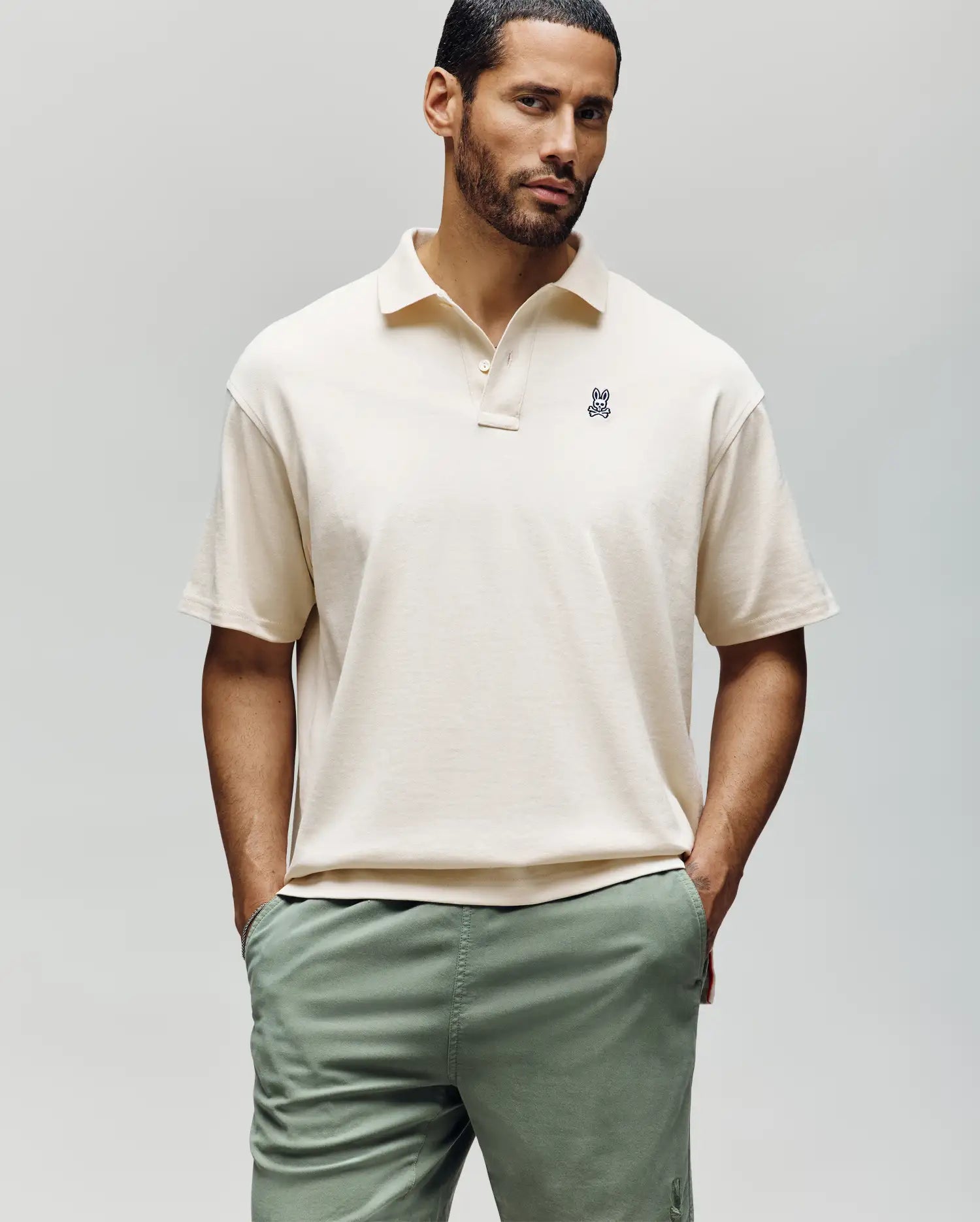 A man stands with hands in pockets, wearing the Psycho Bunny MENS HENRY OVERSIZED FIT POLO - MT0100104 in beige, made from soft Pima cotton jersey with a small bunny logo on the chest, paired with light green pants against a plain background.