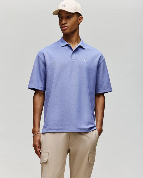 A man wears the Psycho Bunny MENS BENNY OVERSIZED FIT PIQUE POLO - MT0100105 in light blue, paired with beige cargo pants and a beige cap. He stands against a plain background, one hand in his pocket, looking slightly to the side.