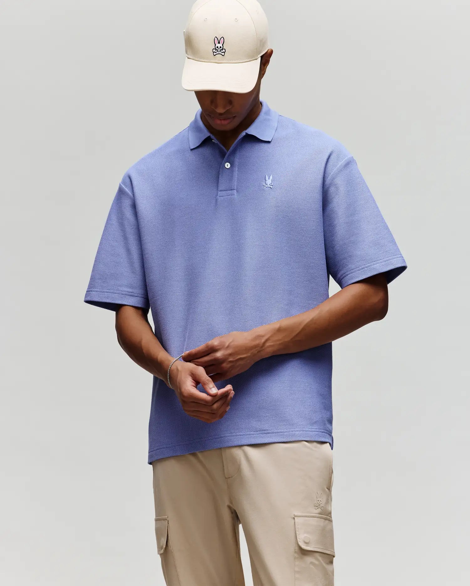 A person wears Psycho Bunny’s MENS BENNY OVERSIZED FIT PIQUE POLO in light purple, beige cargo pants, and a beige cap with the bunny logo, standing against a plain gray background while looking down and adjusting their sleeve.