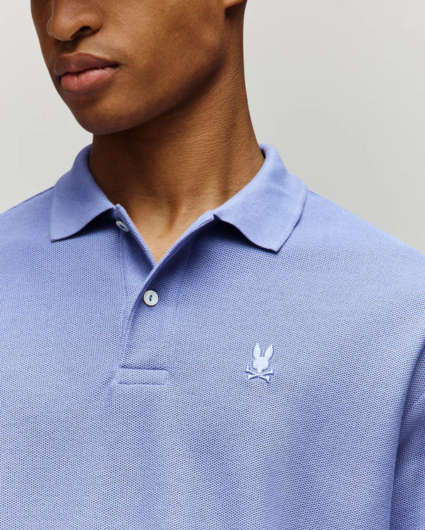 A person wears the Psycho Bunny MENS BENNY OVERSIZED FIT PIQUE POLO - MT0100105 in light blue, featuring a small embroidered rabbit head logo on the left chest. Made of Pima cotton in Peru, the relaxed fit shirt is shown cropped to the face and upper torso.