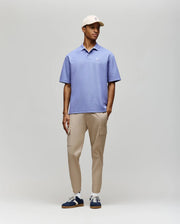 A man wearing the Psycho Bunny MENS BENNY Oversized Fit Pique Polo (MT0100105) in light purple, paired with beige cargo pants, navy and white sneakers, and a beige cap stands against a plain light gray background, looking slightly to the side.