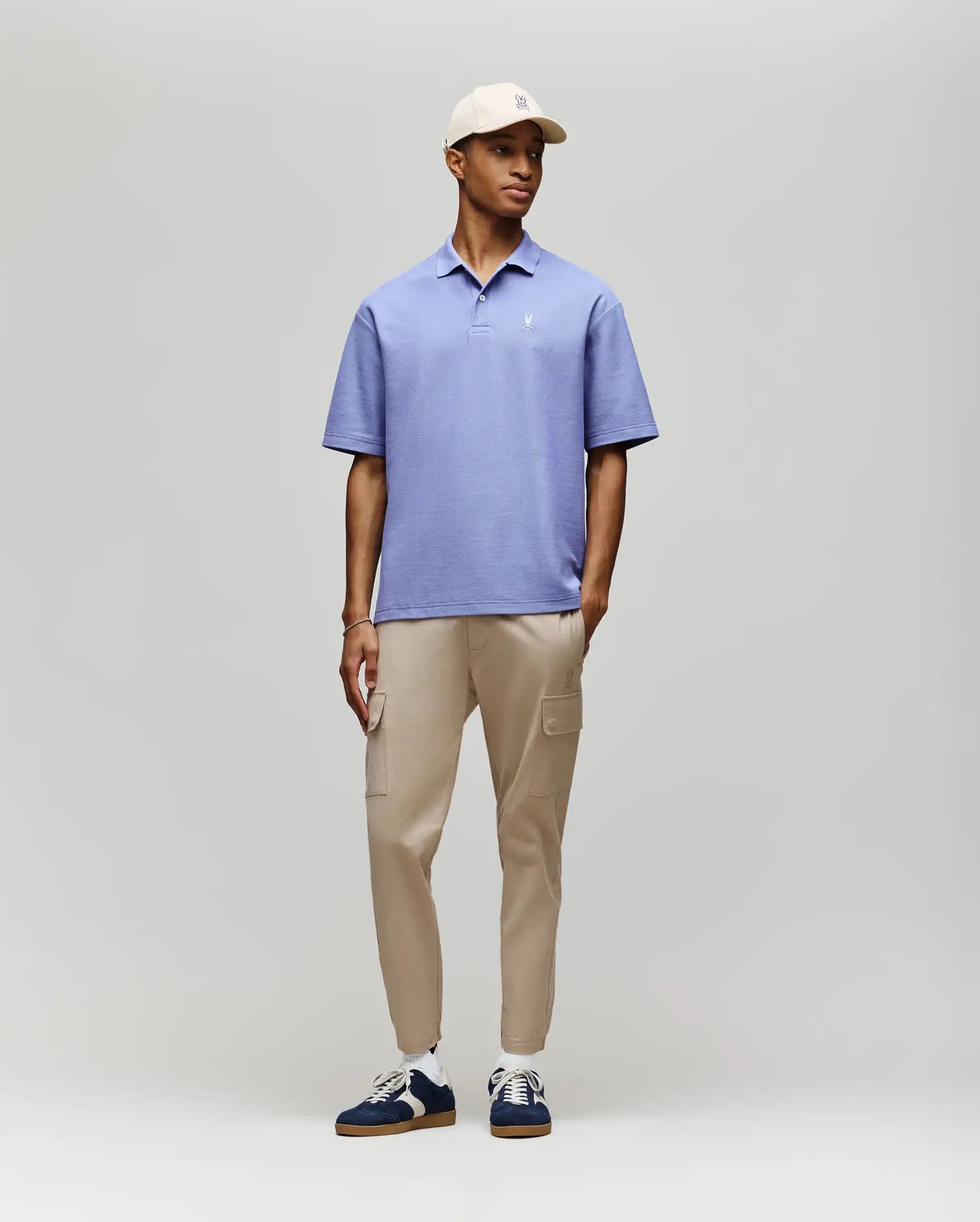 A man wearing the Psycho Bunny MENS BENNY Oversized Fit Pique Polo (MT0100105) in light purple, paired with beige cargo pants, navy and white sneakers, and a beige cap stands against a plain light gray background, looking slightly to the side.