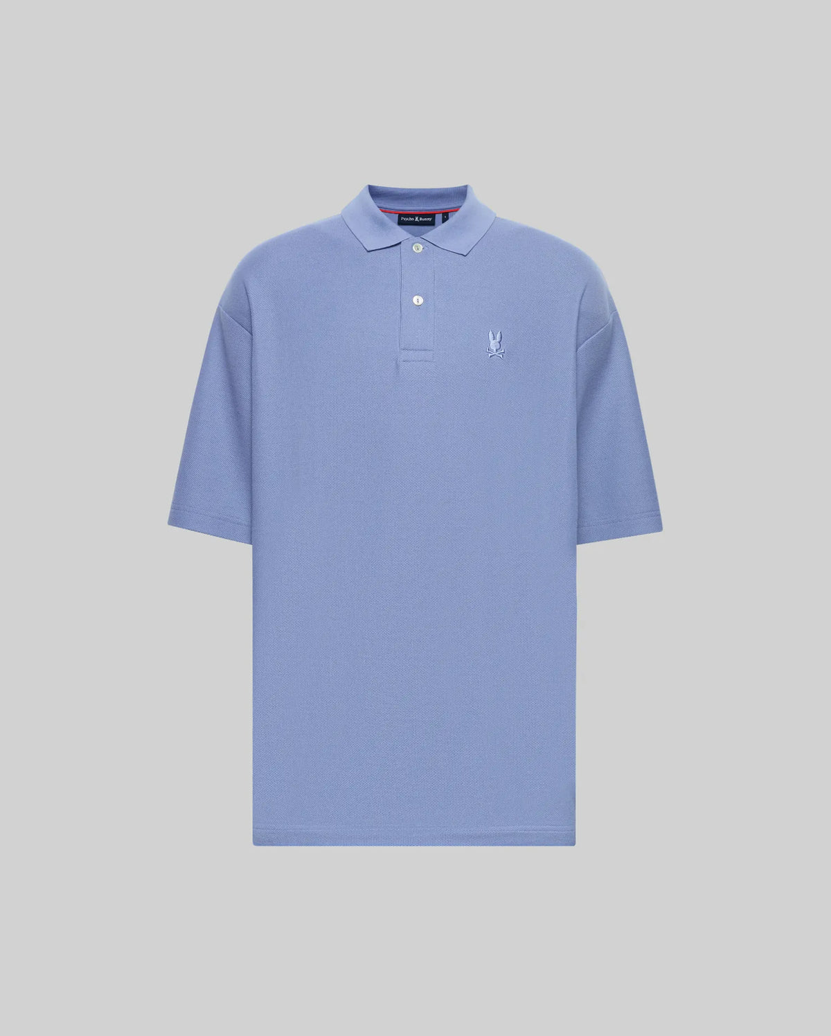The Psycho Bunny MENS BENNY OVERSIZED FIT PIQUE POLO (MT0100105) is a light blue Pima cotton shirt with a collar, two-button neck, relaxed fit, and small white embroidered logo on the left chest. Displayed on a plain light grey background.