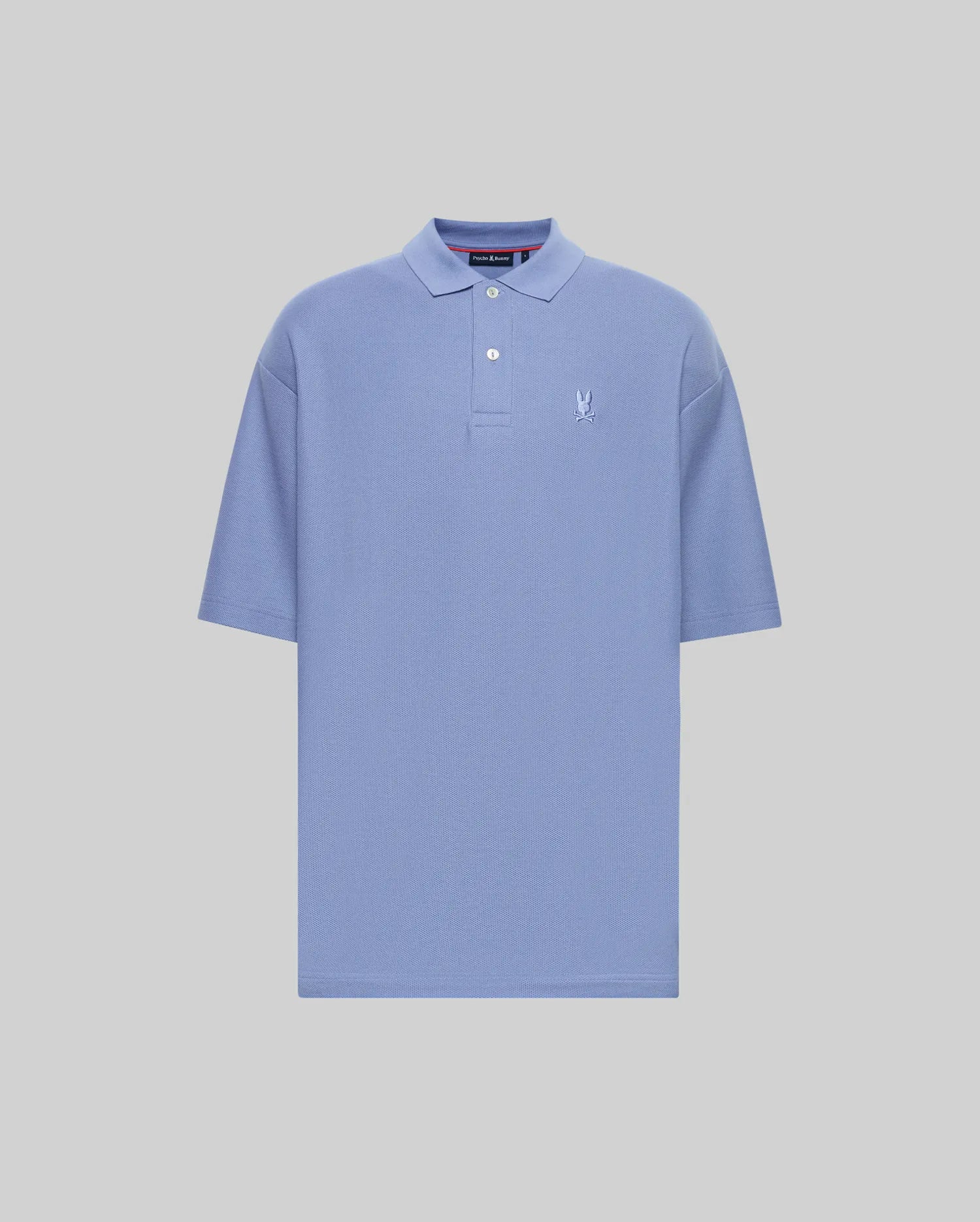 The Psycho Bunny MENS BENNY OVERSIZED FIT PIQUE POLO (MT0100105) is a light blue Pima cotton shirt with a collar, two-button neck, relaxed fit, and small white embroidered logo on the left chest. Displayed on a plain light grey background.