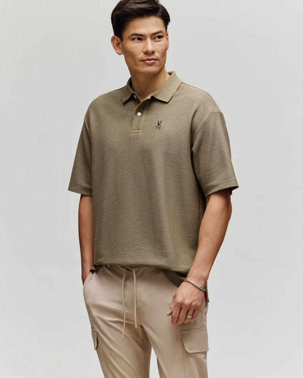 A man wears the Psycho Bunny MENS BENNY OVERSIZED FIT PIQUE POLO (MT0100105) with beige cargo pants, standing with one hand in his pocket and looking slightly to the side against a plain light background.
