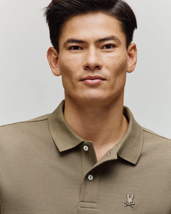 A man with short dark hair wears the Psycho Bunny MENS BENNY OVERSIZED FIT PIQUE POLO (MT0100105), a relaxed light brown polo with a small embroidered rabbit logo, confidently facing the camera against a plain light background.