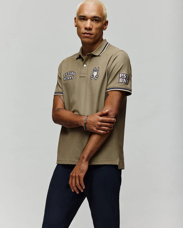 A person with short blond hair wears Psycho Bunny's MENS WADE PATCH PIQUE POLO SHIRT - MT0100109 in khaki, with embroidered Bunny logos and navy pants, standing against a light background, looking at the camera with arms crossed.