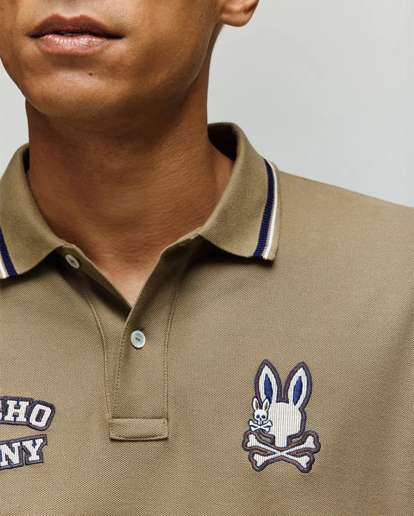 A close-up of a person wearing the Psycho Bunny MENS WADE PATCH PIQUE POLO SHIRT in khaki, with navy and white collar trim and an embroidered Bunny above crossed bones on the left chest.